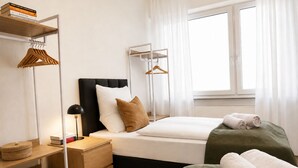 Een moderne slaapkamer met een bed, nachtkastjes, een bedlampje, een raam met gordijnen en een plank met boeken en kledinghangers.