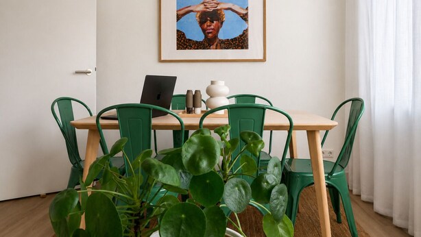 Een eethoek met een tafel, stoelen, een laptop, een lijst met een afbeelding en een potplant.