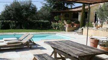 A pool area with lounge chairs and a wooden table, surrounded by greenery and a building with a tiled roof.