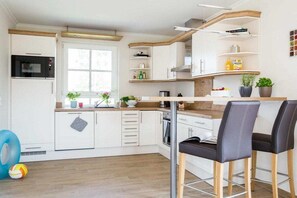 A modern kitchen with white cabinets, a built-in oven, and a bar area with two chairs. - Holiday home Reed Warbler 43 (Fuhlendorf)