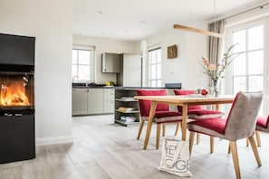 A modern living room with a fireplace, a dining area with red chairs, and a kitchen in the background. - Holiday home with 1 bedroom (Fuhlendorf)