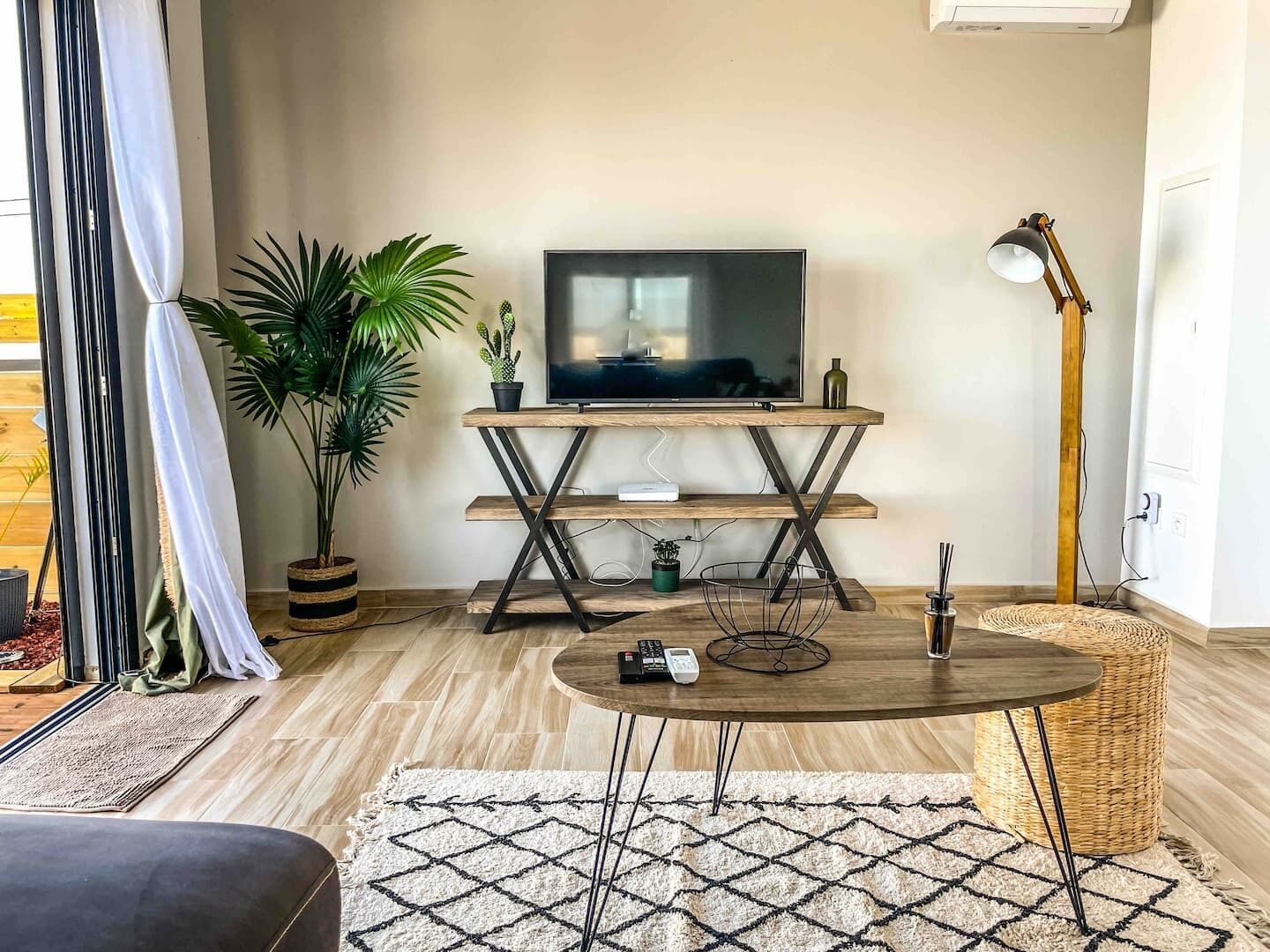 Una sala de estar con un televisor sobre un mueble de madera, una mesa redonda de centro, una lámpara de pie y plantas en macetas.