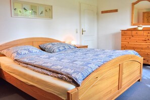 A wooden bed with a patterned bedspread and pillows, a bedside table with a lamp, a wooden dresser, and a framed bird illustration on the wall. - Knut in the Mill Park (Prerow)