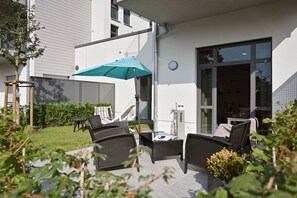 An outdoor seating area with wicker furniture and a blue umbrella, adjacent to a building with large windows and a door. - Holiday apartment in Glowe with swimming pool (Glowe)