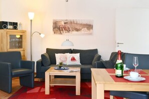 Ein Wohnzimmer mit einer Couch, zwei Sesseln, einem Couchtisch, einer Lampe, einem Gemälde, einem Schrank und einem Tisch mit Flasche und Glas.