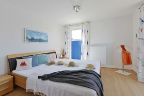 Ett sovrum med en säng, sängbord, ett litet bord, ett fönster med gardiner, en tavla på väggen och en stol med en handduk.