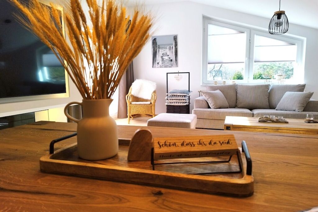 Un salon avec une table en bois, un vase contenant des plantes séchées, une chaise en osier, un canapé et un tableau encadré accroché au mur.