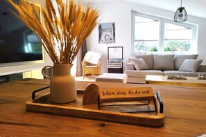 Uma sala de estar com uma mesa de madeira, um vaso com plantas secas, uma cadeira de vime, um sofá e um quadro na parede.