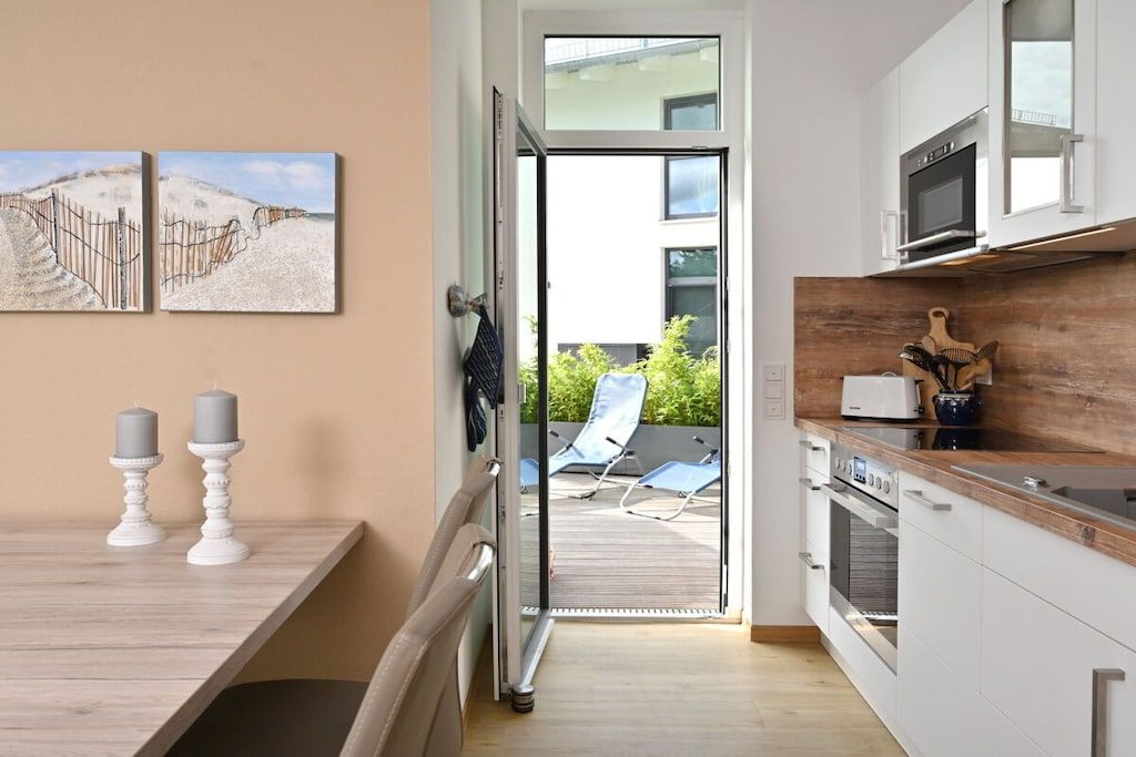 Une cuisine moderne dotée d’un plan de travail en bois, d’un four à micro-ondes et d’un four traditionnel. Une porte-fenêtre coulissante donne accès à un espace extérieur comprenant une terrasse et des chaises longues.