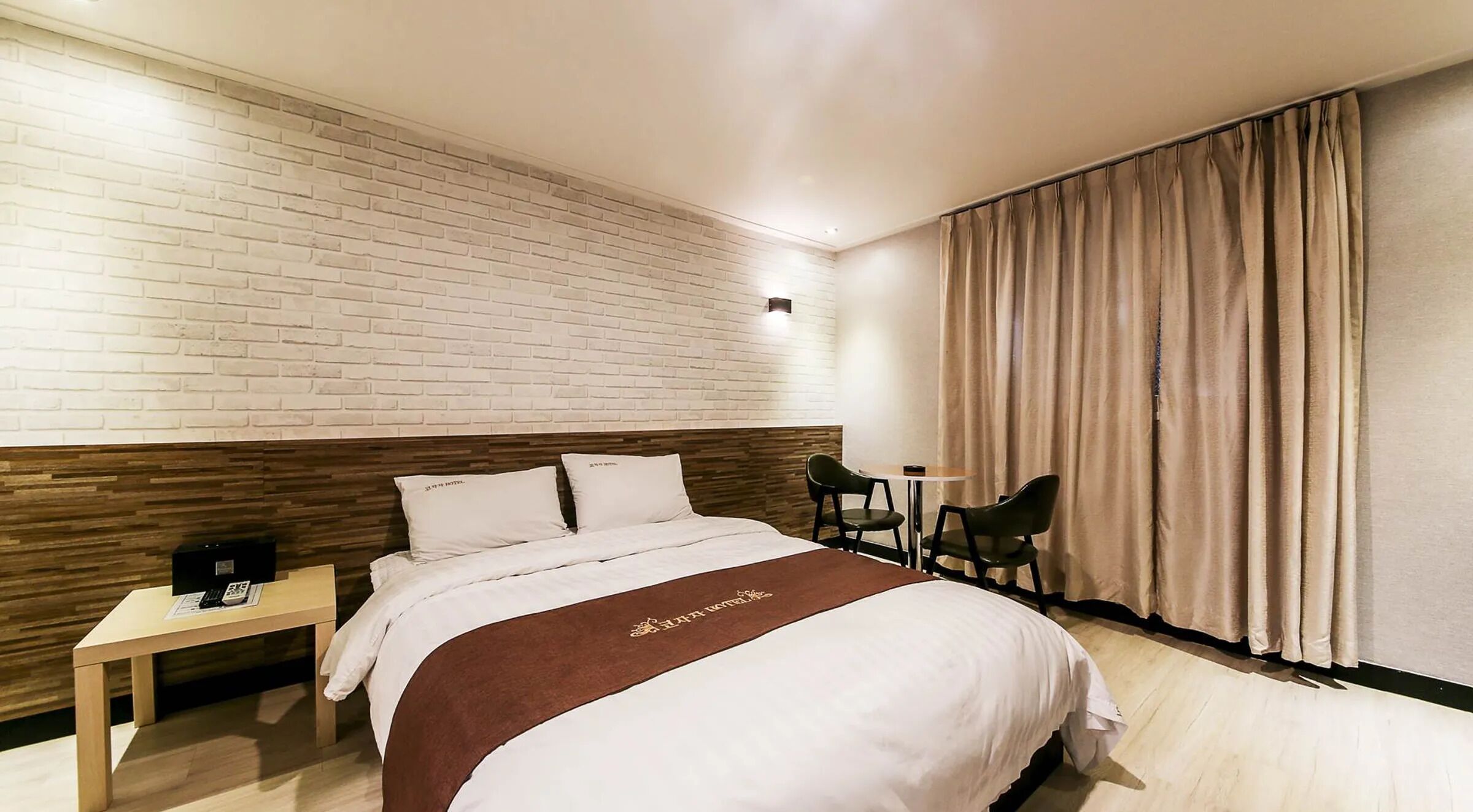 Camera d'albergo con parete a effetto mattone, una testiera in legno, un letto con biancheria bianca, una coperta marrone, un comodino con un telefono e una finestra con tende beige.