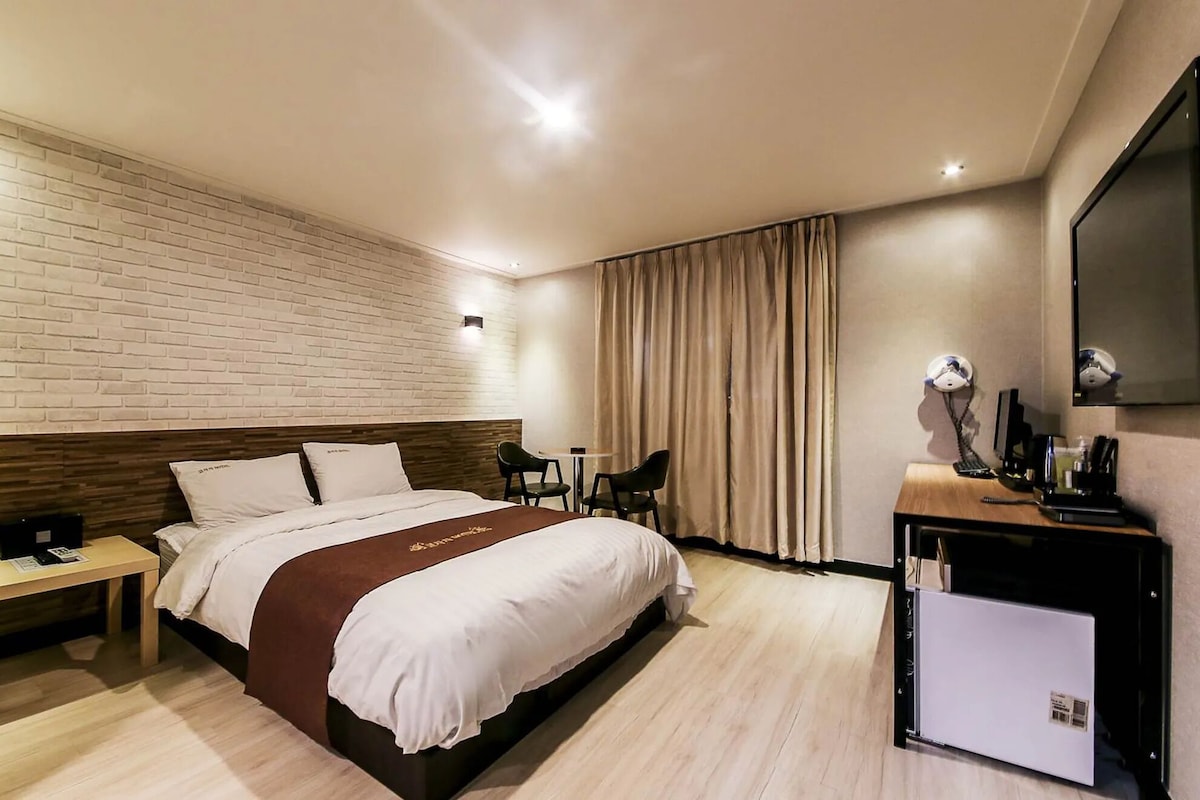 A hotel room with a bed, bedside table, desk, chair, TV, and a minibar.