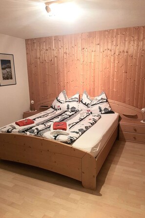 A room with a wooden bed, a wooden headboard, a nightstand, and a wall-mounted picture. - Ferienwohnungen Teuriblick A&B, (Splügen), by Interhome (Splügen)