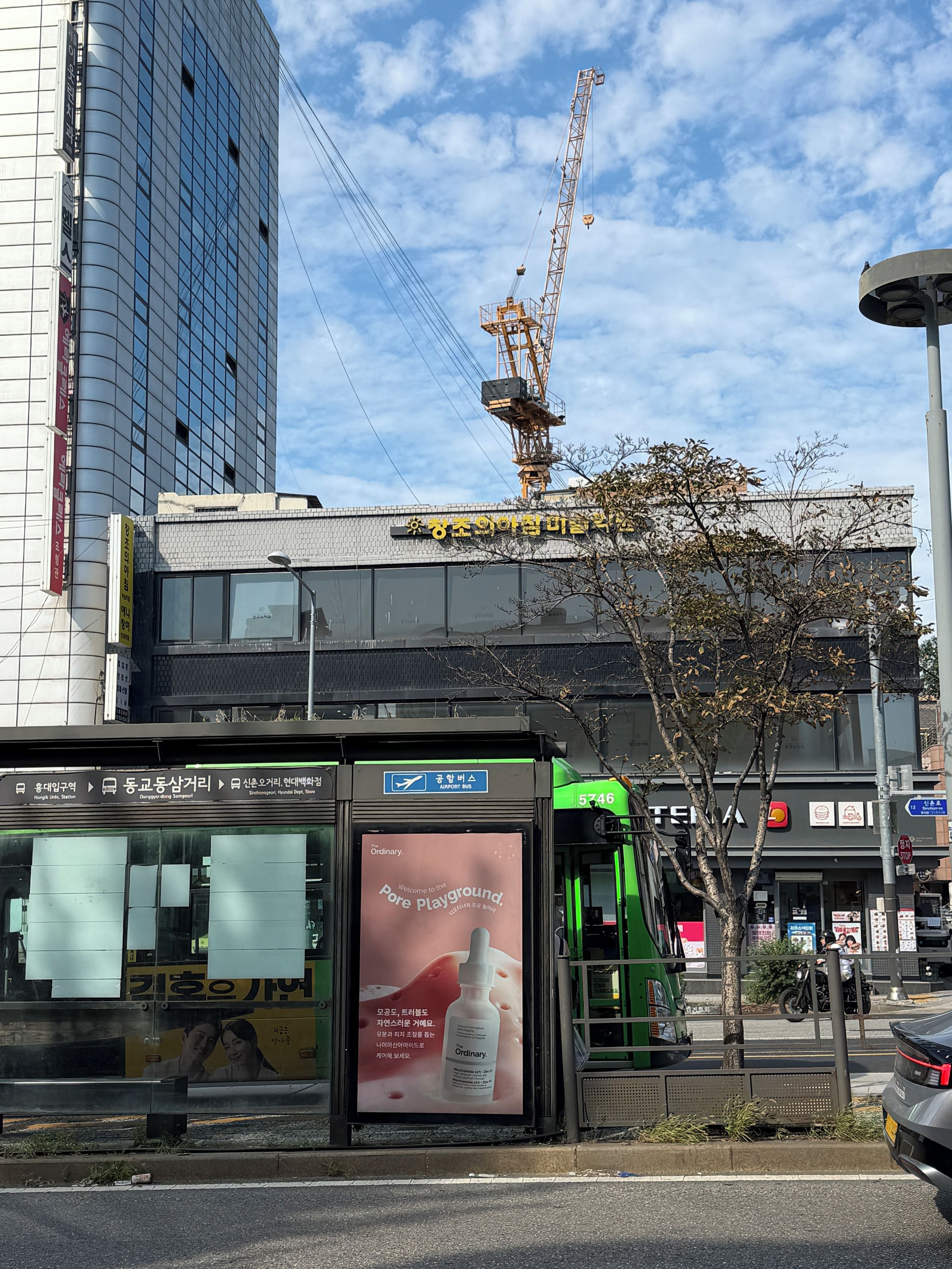 A bus stop with a sign for the airport bus and a billboard advertising a skincare product.