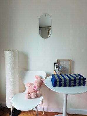 A room with a white chair, a small table with a blue and white striped cloth, a pink stuffed toy, a white lamp, and a framed picture on the wall.