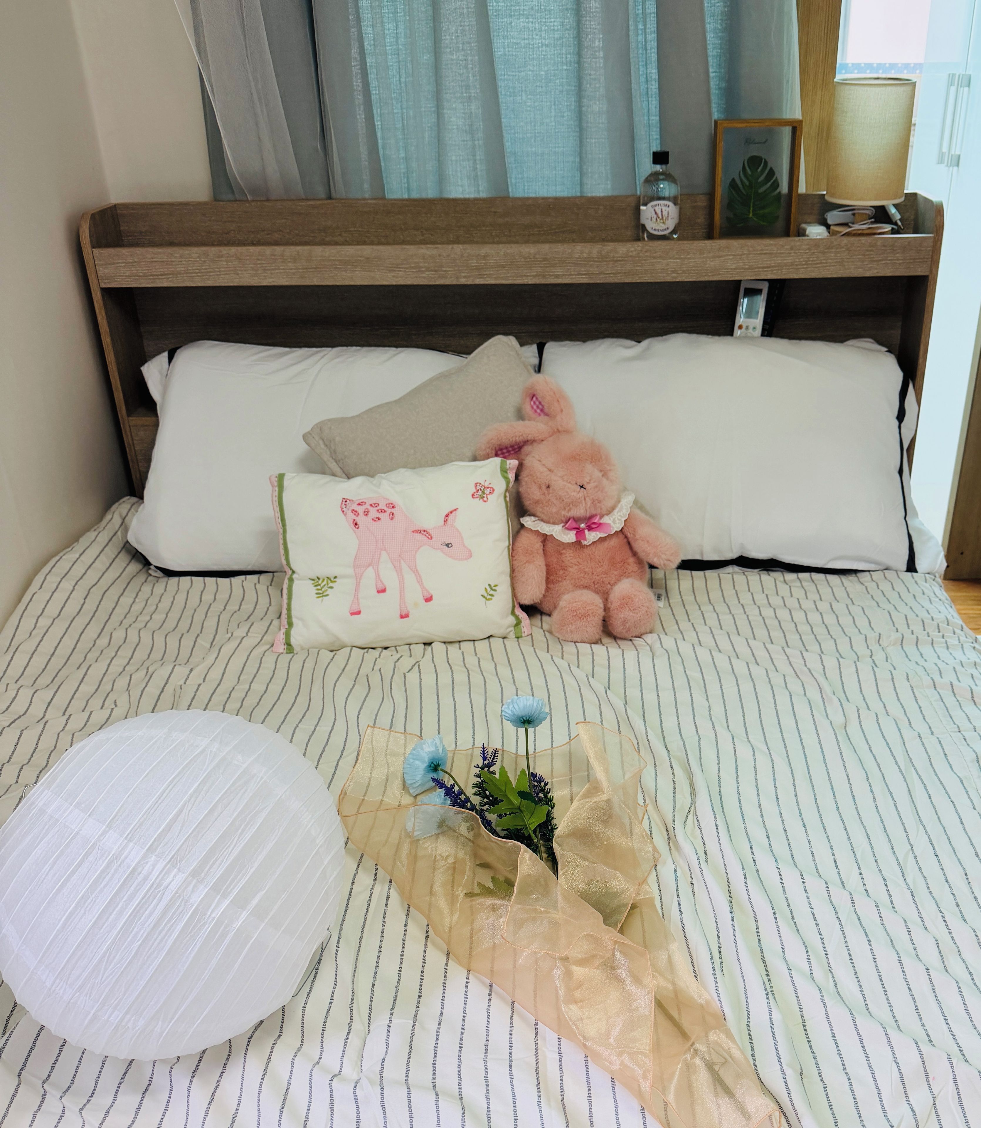 A bed with a striped bedspread, a pillow with a cow design, a teddy bear, and a small bouquet of flowers.