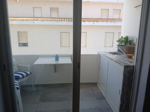 Un pequeño balcón con una mesa, una silla, una planta en maceta y vistas a un edificio con ventanas y tejado de tejas.