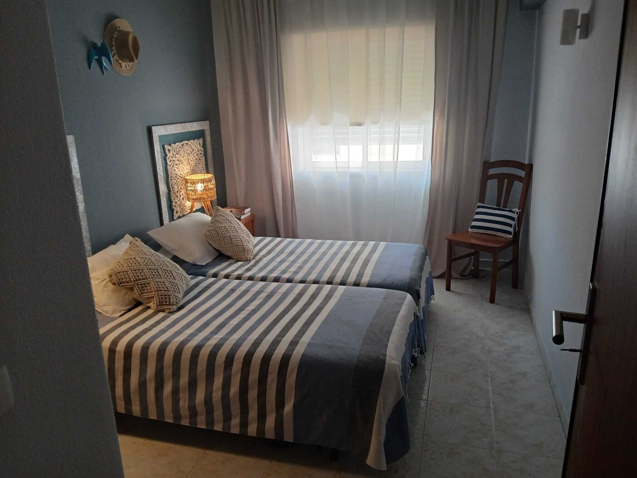 Un dormitorio con dos camas, una mesita de noche, una silla, un adorno en la pared y una ventana con cortinas.