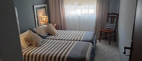 Un dormitorio con dos camas, una mesita de noche, una silla, un adorno en la pared y una ventana con cortinas.