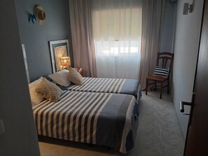 A bedroom with two beds, a nightstand, a chair, a decorative wall piece, and a window with curtains. - Pé na Areia House, Armação de Pêra (Armação de Pêra)