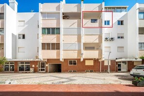 Un edificio residencial de varios pisos con balcones, un patio empedrado y un coche blanco estacionado al frente.