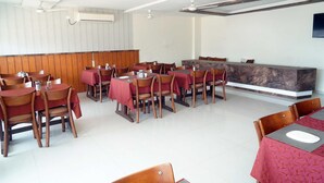 A restaurant with multiple tables and chairs, a wooden panel wall, ceiling fans, and a flat-screen TV mounted on the wall. - Deluxe Hotel Utkarsh (Silvassa)