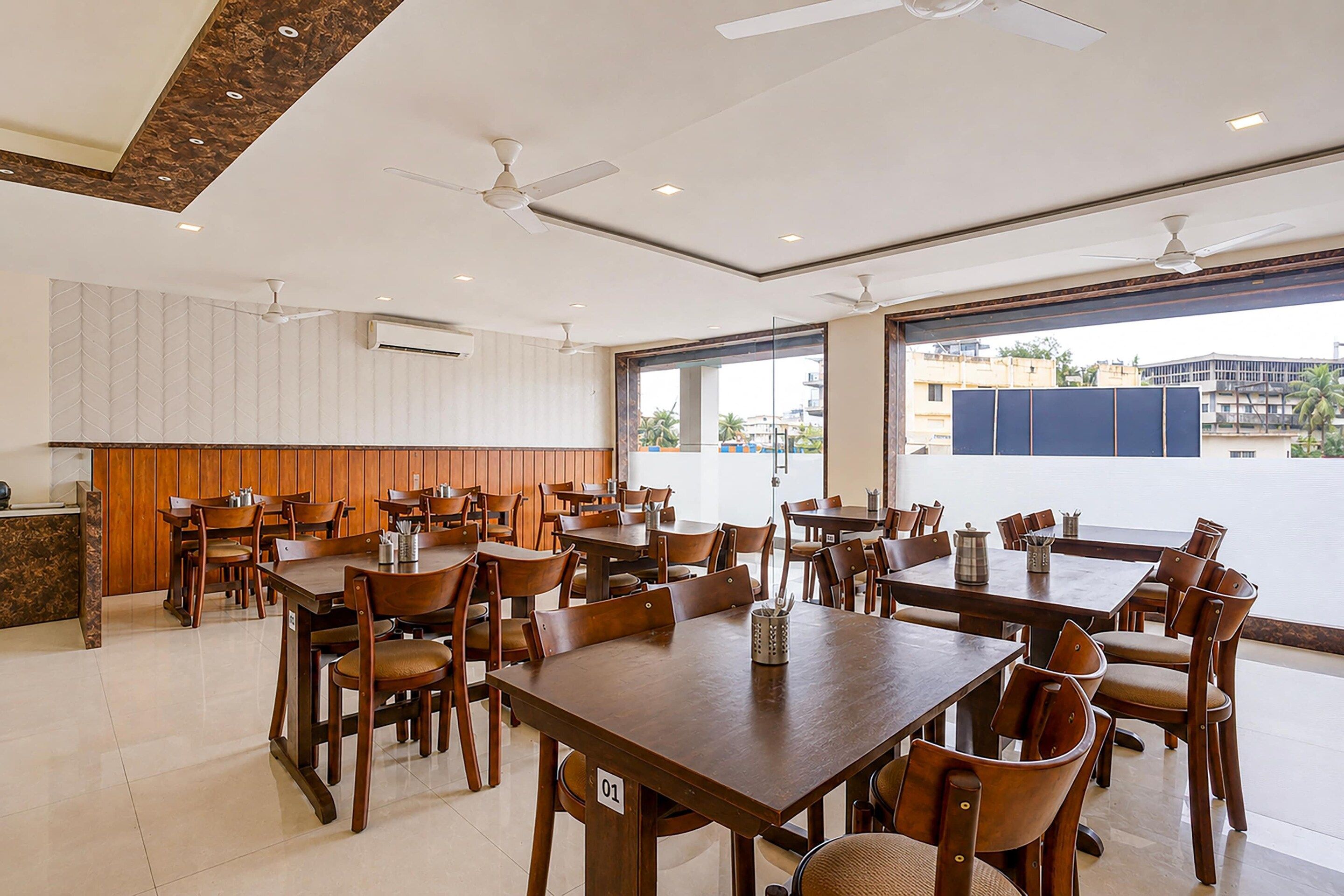 Un comedor amplio con varias mesas y sillas de madera, ventiladores de techo y grandes ventanales que ofrecen vistas al exterior.