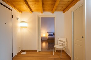 A hallway with a wooden floor, a white chair, a bedside table with a lamp, and a bed in the adjacent room. - Le Residenze dell'Alloro - Gorizia Home (Palermo)