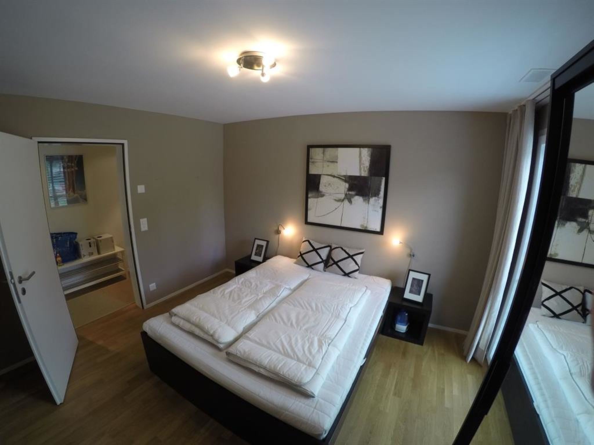 Un dormitorio con cama, mesitas de noche, un cuadro en la pared y una puerta que da a otra habitación.