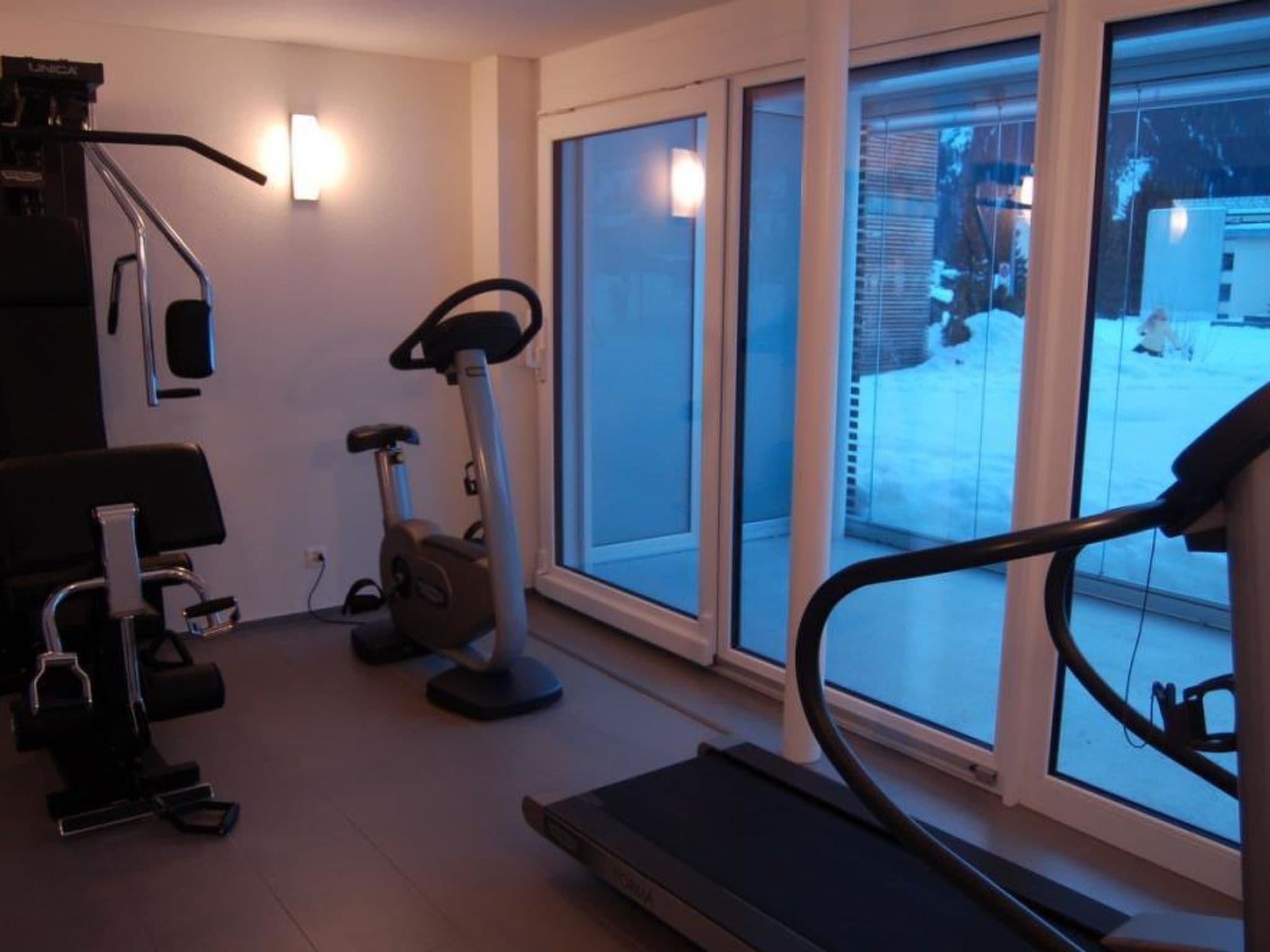 Un gimnasio en casa con equipo de ejercicio, incluyendo una cinta de correr, bicicleta estática y elíptica, con ventanas amplias que ofrecen vistas al exterior.