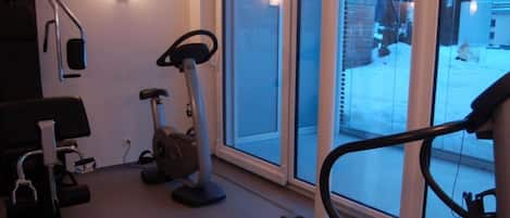 Un gimnasio en casa con equipo de ejercicio, incluyendo una cinta de correr, bicicleta estática y elíptica, con ventanas amplias que ofrecen vistas al exterior.