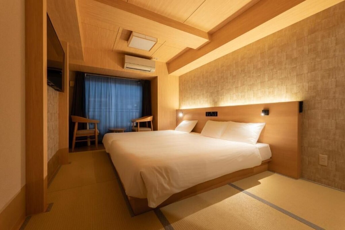 A hotel room with a large bed, a wooden headboard, a small seating area, and a ceiling-mounted air conditioning unit.