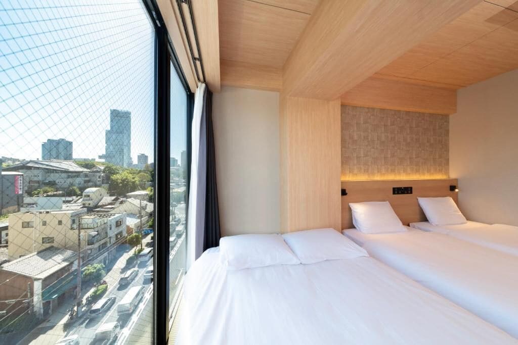 Une chambre d’hôtel dotée d’une grande fenêtre, d’un lit avec du linge de maison blanc, d’une tête de lit en bois clair et offrant une vue sur le paysage urbain.