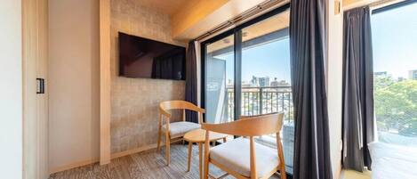 A hotel room with a large window, a flat-screen TV mounted on the wall, and two wooden chairs.