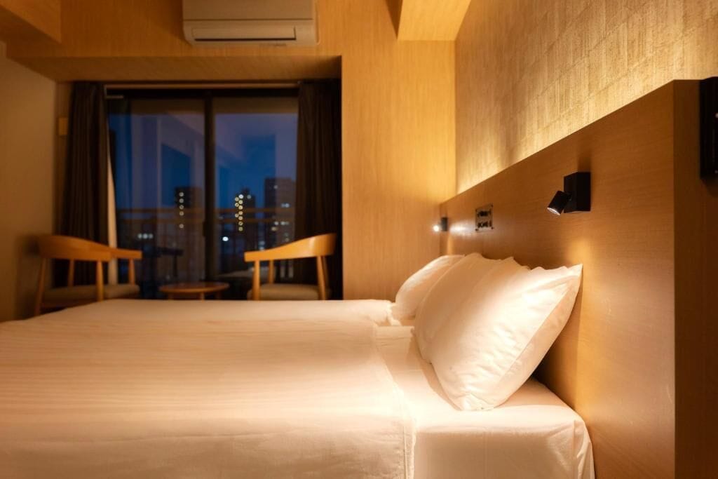 Ein Hotelzimmer mit Bett, kleinem Tisch, zwei Stühlen und Blick auf eine Stadtlandschaft durch ein großes Fenster.