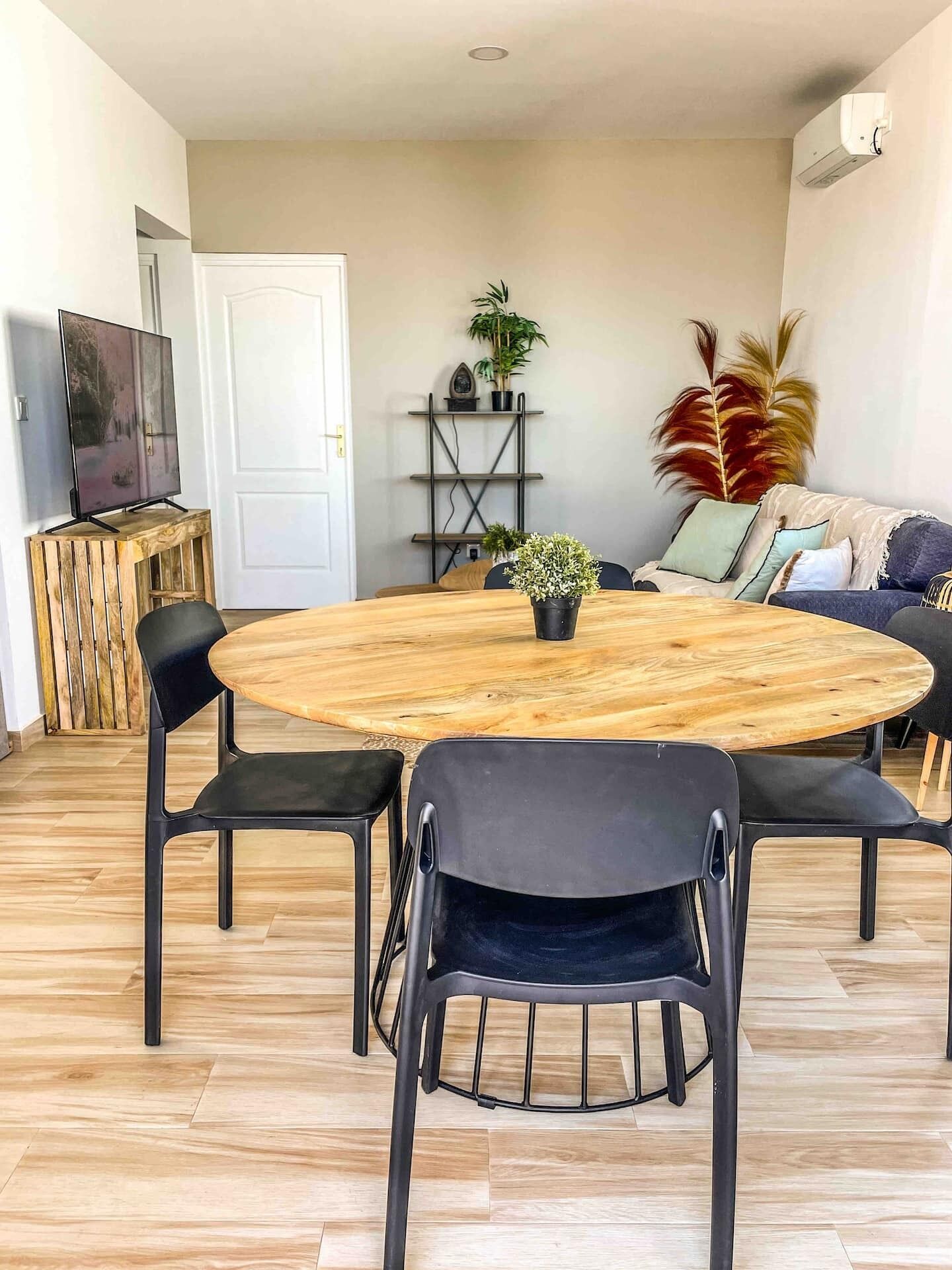 Una mesa redonda de madera con cuatro sillas, una planta en maceta y un televisor sobre un mueble en una habitación con suelo de madera y un sofá.