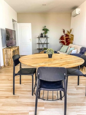 Una mesa redonda de madera con cuatro sillas, una planta en maceta y un televisor sobre un mueble en una habitación con suelo de madera y un sofá.