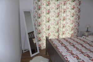 Ein Schlafzimmer mit einem hölzernen Bett, einem weißen, flauschigen Teppich, einem Nachttisch mit Lampe und blumigen Vorhängen.