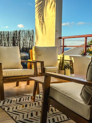 A sunlit outdoor seating area with a white sofa, a wooden table, and a patterned rug.