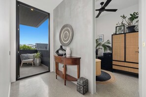 A modern interior with a sliding glass door leading to a balcony, a wooden console table with decorative items, a woven basket, and a traditional cabinet with plants. - Black on Black Wattle - Beachfront, Rooftop, Pool (Casuarina)