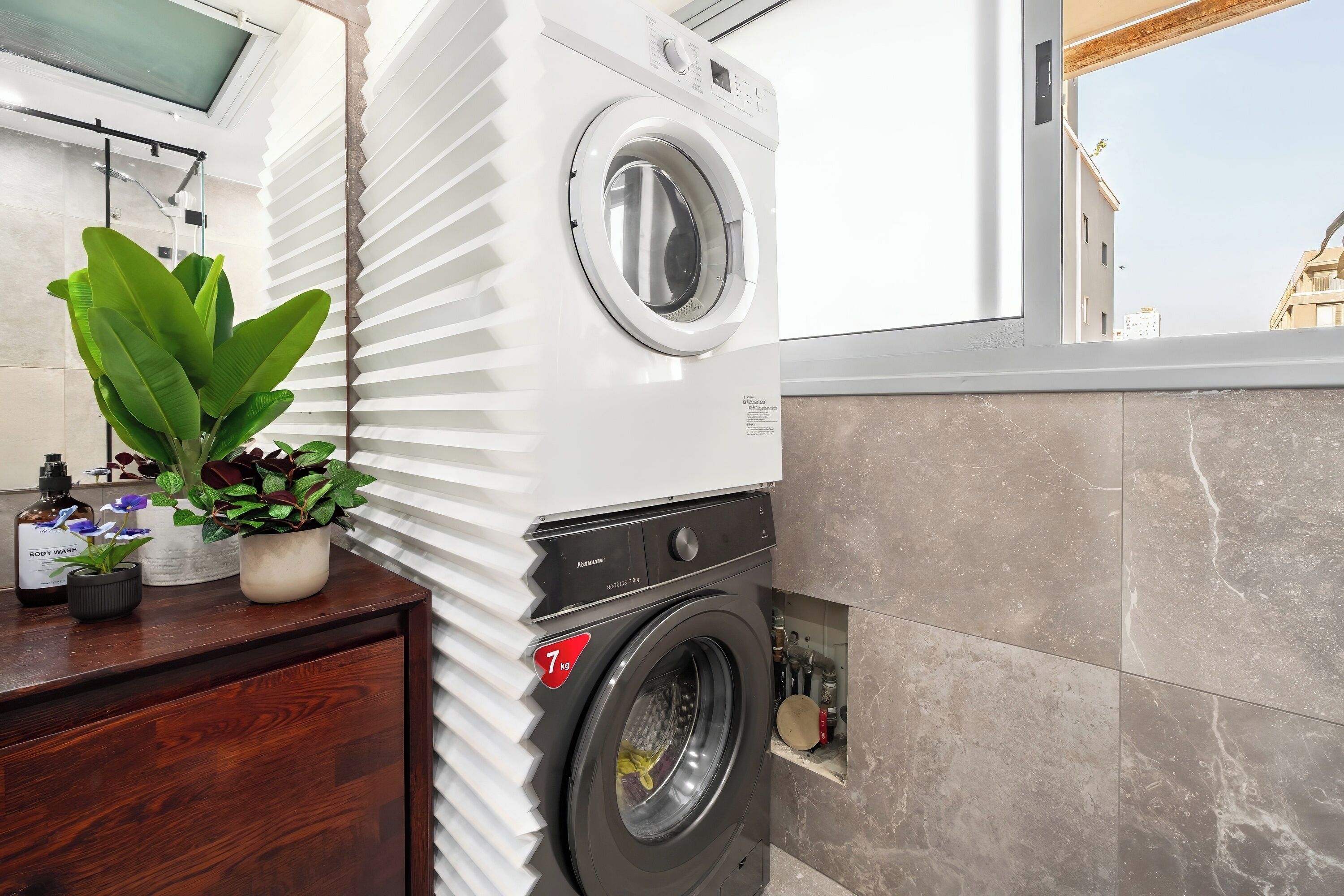 A modern laundry room with a front-loading washing machine, a wooden cabinet with a potted plant, and a view of the outside through a window.