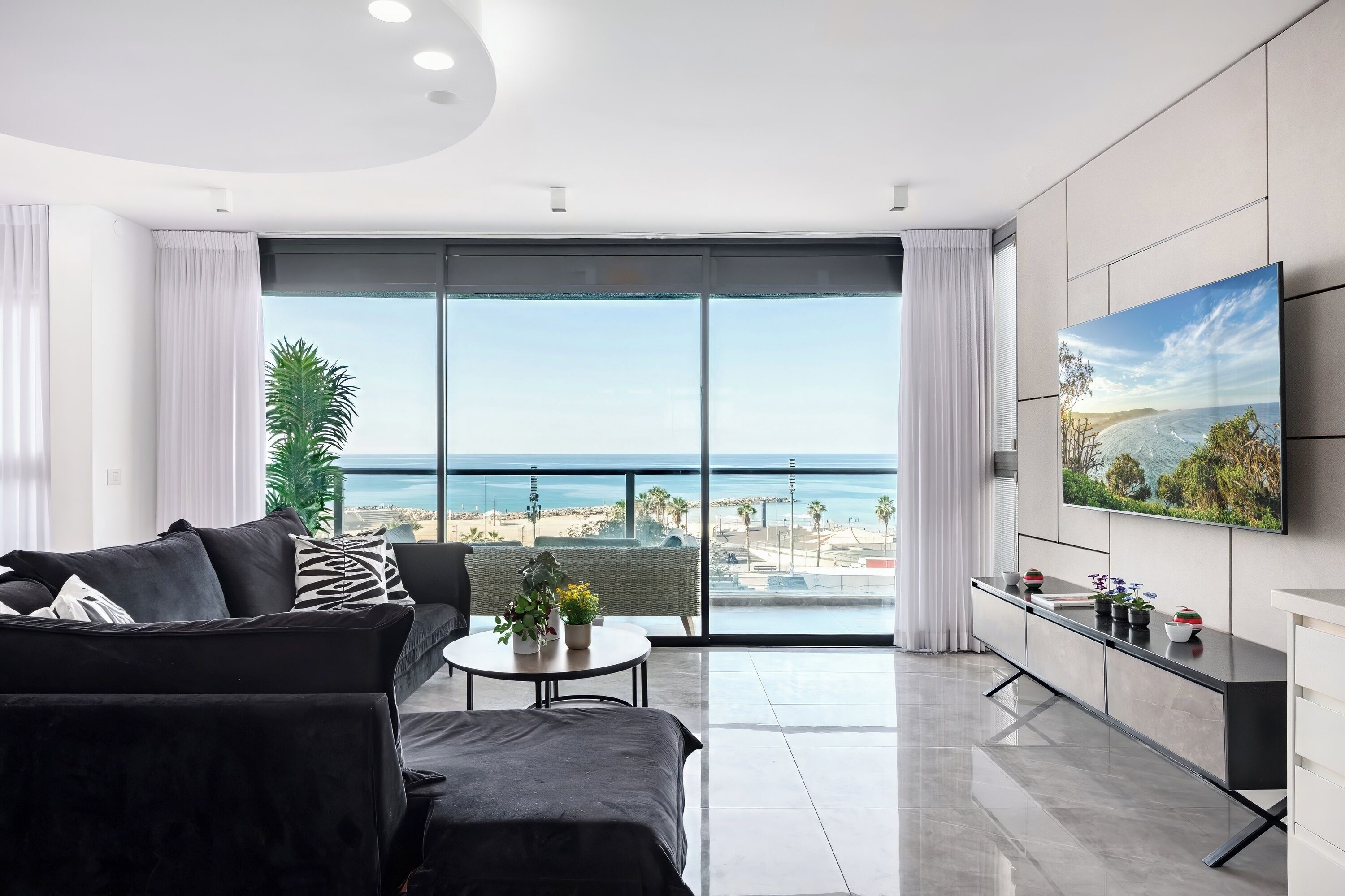 Ein modernes Wohnzimmer mit großem Fernseher, einer Couch, einem Couchtisch und Blick auf den Strand.