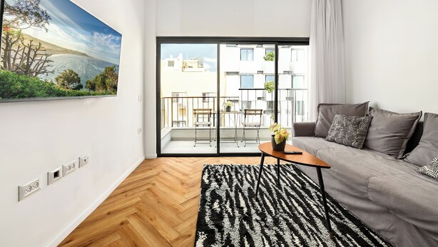 A modern living room with a sofa, a coffee table, and a view of a balcony with outdoor furniture. - Stylish 2BR City Apartment Near the Beach & Bograshov (Tel Aviv-Yafo)