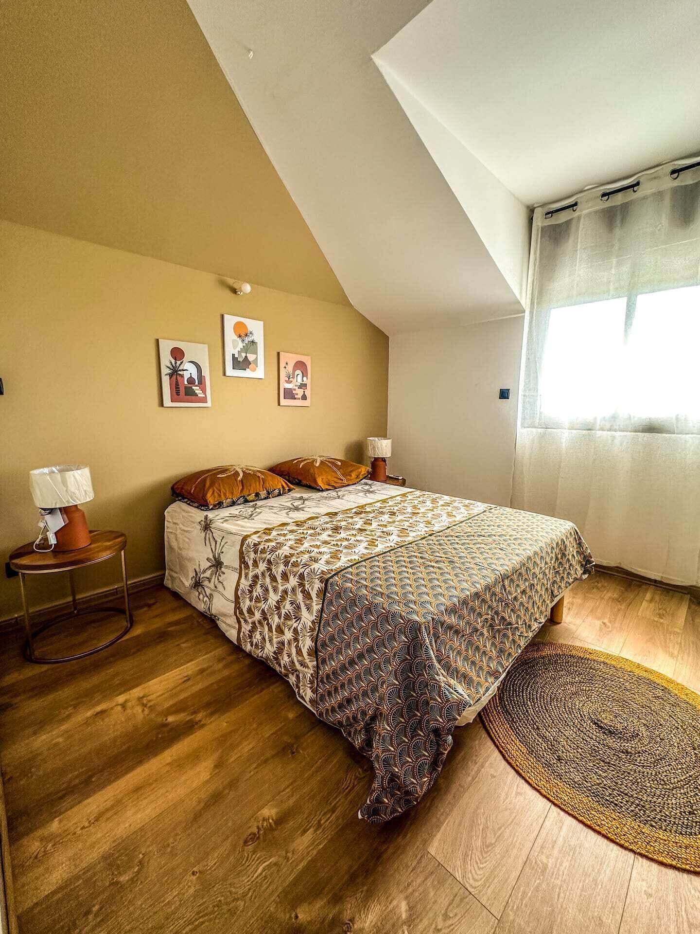 A bedroom with a sloped ceiling, a bed with patterned bedding, a small bedside table with a lamp, and a window with curtains.