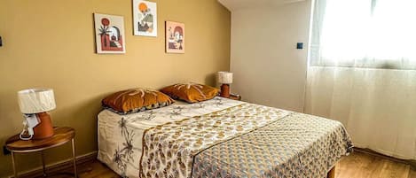 A bedroom with a sloped ceiling, a bed with patterned bedding, a small bedside table with a lamp, and a window with curtains.