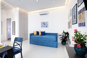 Ein modernes Wohnzimmer mit einem blauen Sofa, einem Sessel und einem Esstisch. An den Wänden sind Klimaanlagen montiert und es gibt gerahmte Kunstwerke.