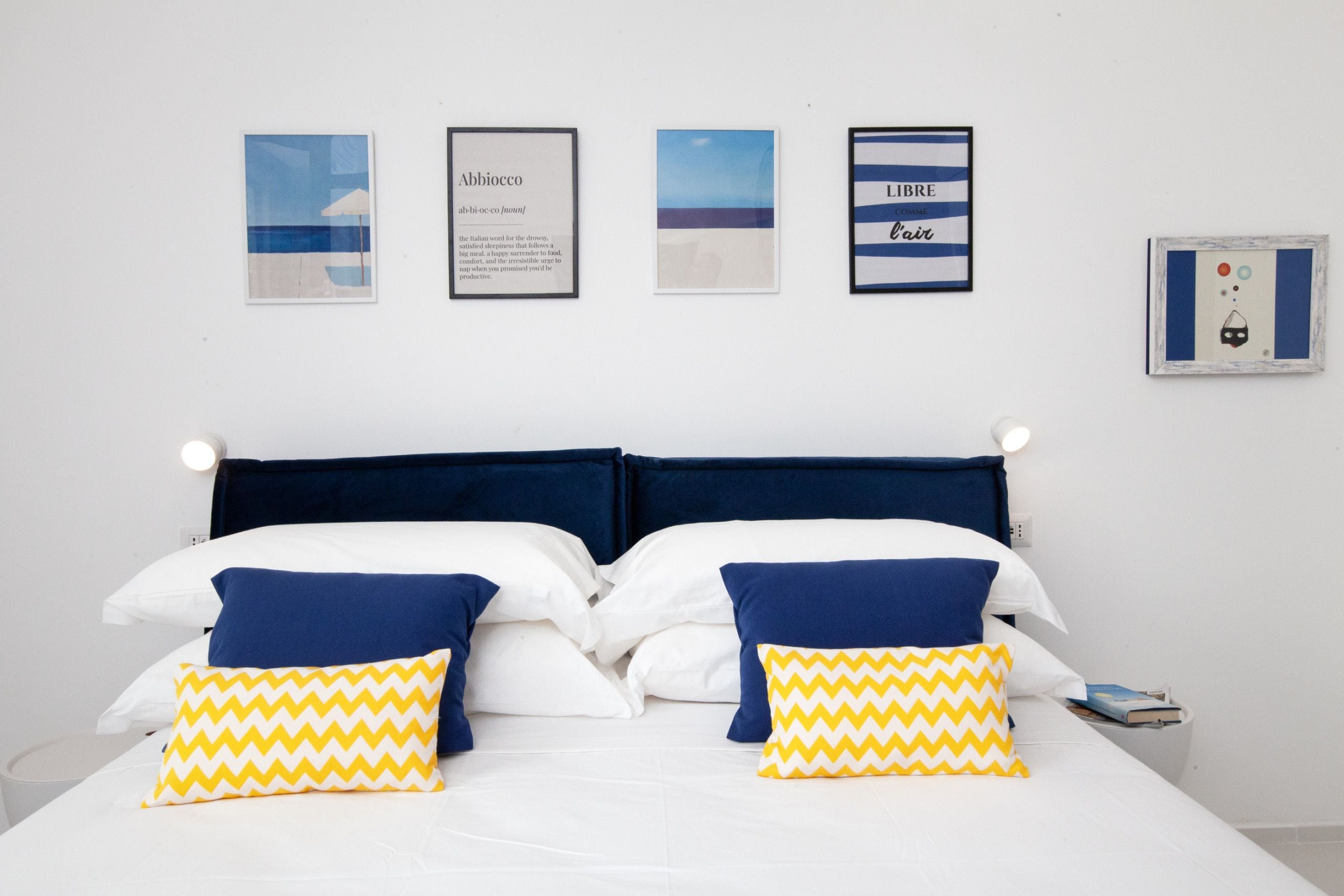 Un lit avec deux oreillers, une table de chevet sur laquelle se trouve un livre, et quatre tableaux encadrés accrochés au mur.