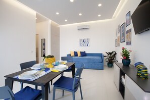 Ein modernes Wohnzimmer mit einem blauen Sofa, einem Esstisch mit Tellern und Gläsern sowie einer Küchenzeile mit Fernseher und Topfpflanzen.