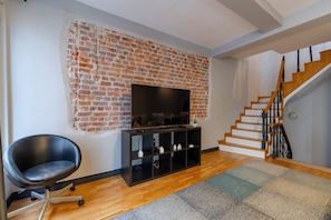A living room with a brick wall, a flat-screen TV, a black chair, a wooden staircase, and a patterned rug.