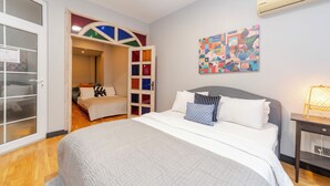 Ein Schlafzimmer mit einem Bett, einem Nachttisch, einer Wand-Klimaanlage und einem farbenfrohen abstrakten Gemälde.