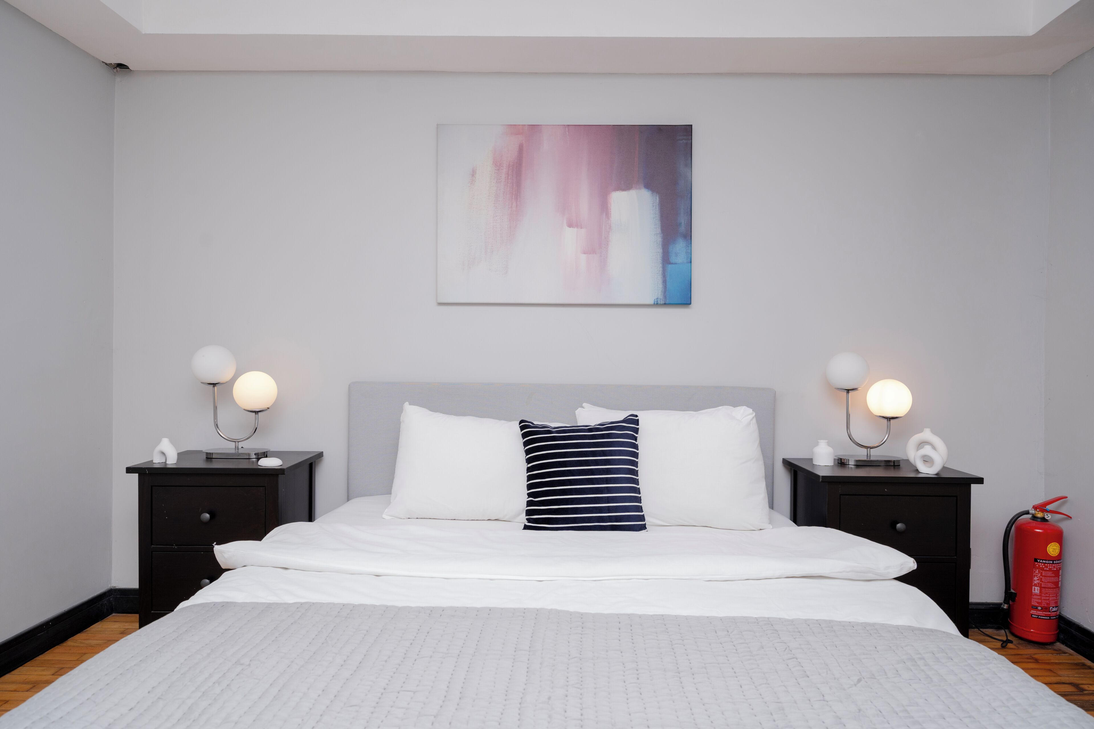 A neatly arranged bedroom with a large bed, two bedside tables with lamps, a fire extinguisher, and a wall-mounted abstract painting.